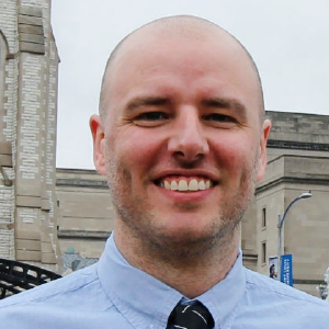 A photo of Evan Rhinesmith. He has a smile that involves his eyes and dimples appear. He is a clean shaven bald man standing in front of some sort of museum.